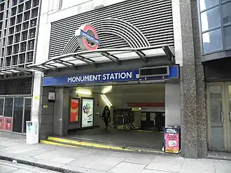 An entrance in a larger building under a sign reading "MONUMENT STATION" reveals banisters leading down. A woman is walking out of the entrance